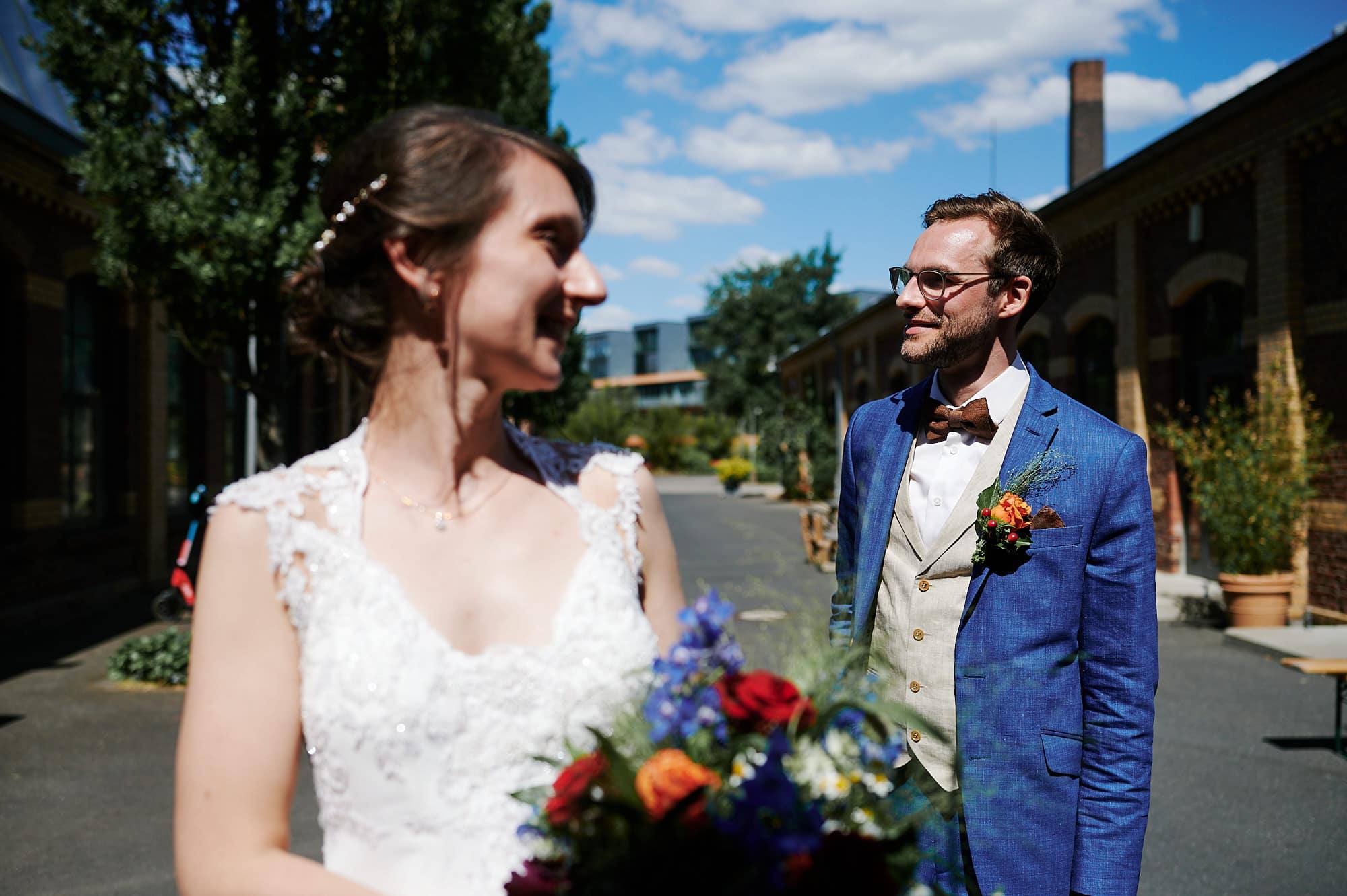 Eine Braut und ein Bräutigam lächeln sich an einem sonnigen Tag im Freien an, während der Bräutigam die Braut bewundernd ansieht.