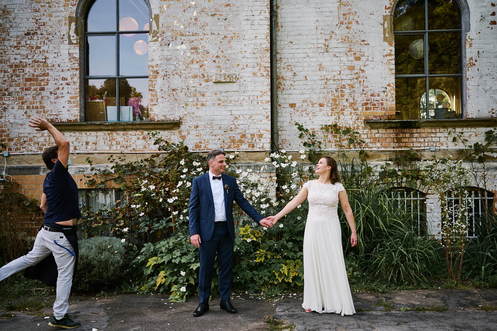 Der Hochzeitsfotograf fängt einen offenen Moment ein, in dem ein fröhliches Brautpaar Händchen haltend vor einer rustikalen Backsteinmauer.