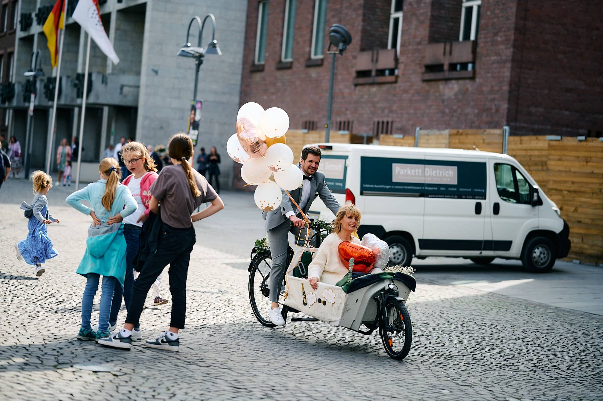 Ein Brautpaar mit Luftballons fährt auf einem Lastenfahrrad, während Fußgänger vorbeigehen.