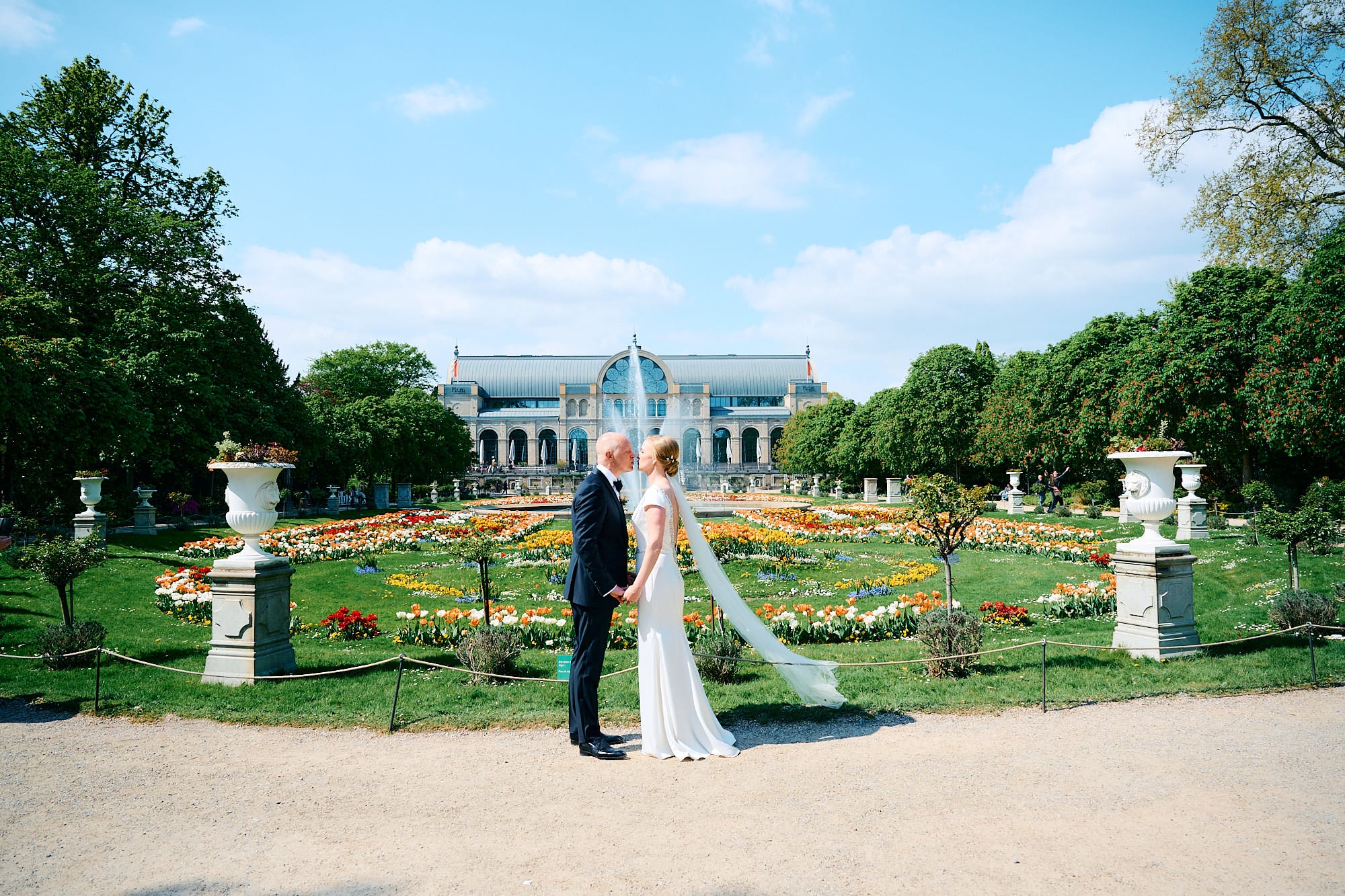 Händchen haltendes Paar in Hochzeitskleidung in einem formellen Garten mit einem klassischen Gebäude im Hintergrund.
