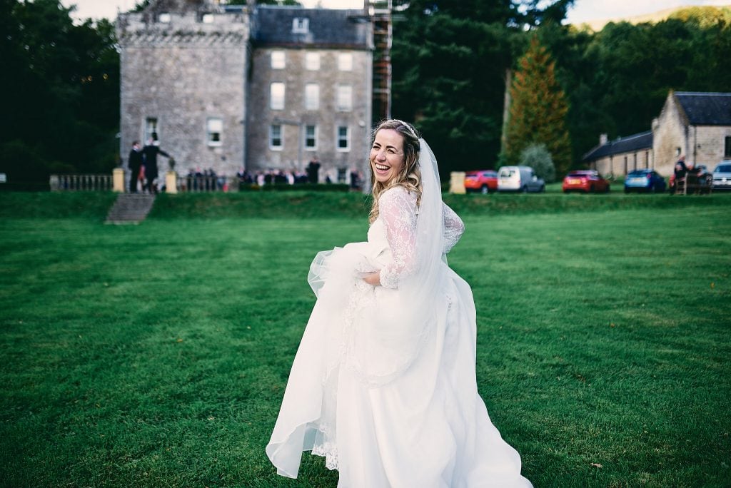 Eine fröhliche Braut in einem weißen Kleid und Schleier blickt auf einer Rasenfläche von Culcreuch Castle in Schottland über ihre Schulter, mit dem historischen Gebäude im Hintergrund.