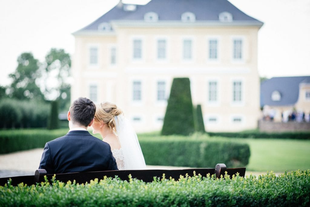 Das Brautpaar auf einer Bank mit dem Schloss im Hintergrund.