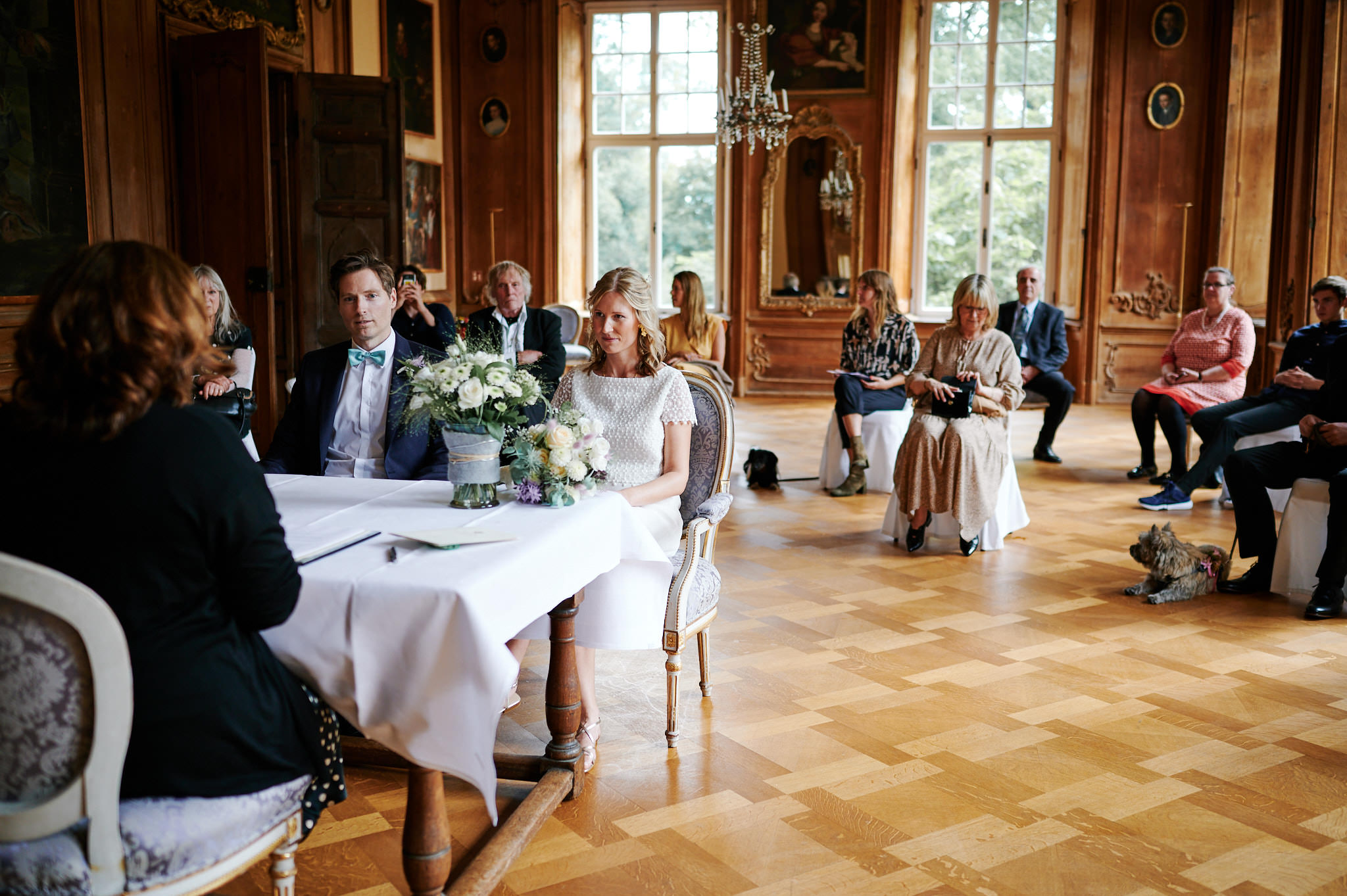 Ein Paar unterzeichnet Dokumente bei seiner Hochzeitszeremonie in einem kunstvoll dekorierten Raum, während die Gäste zuschauen.
