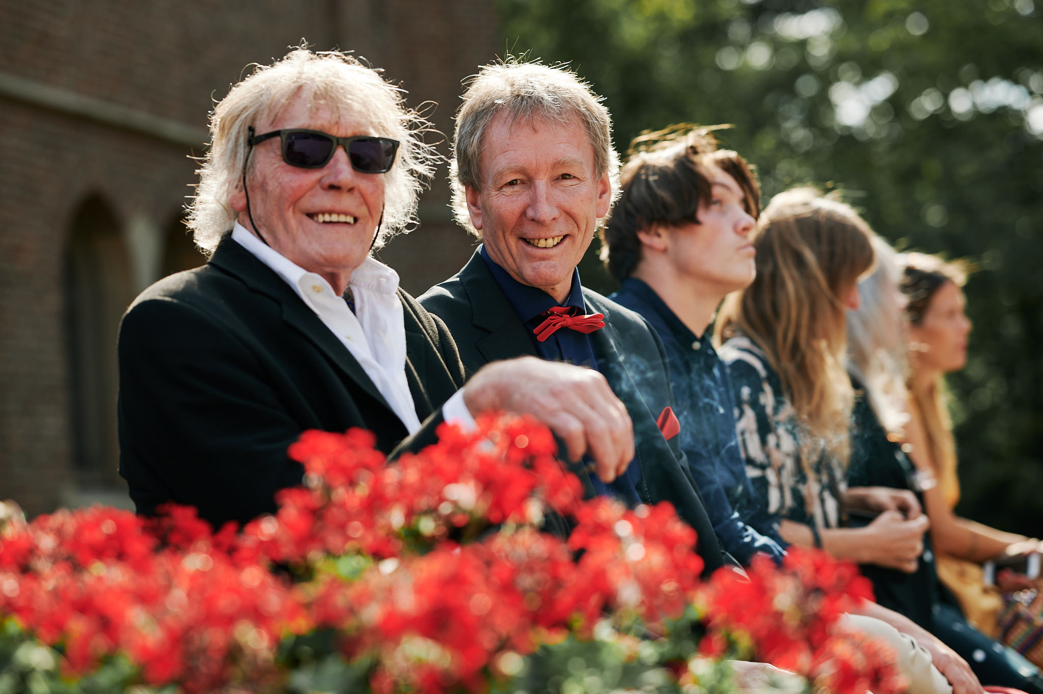 Zwei lächelnde Männer in formeller Kleidung sitzen vor einem Blumenbeet bei einer Outdoor-Veranstaltung auf Schloss Wissen.