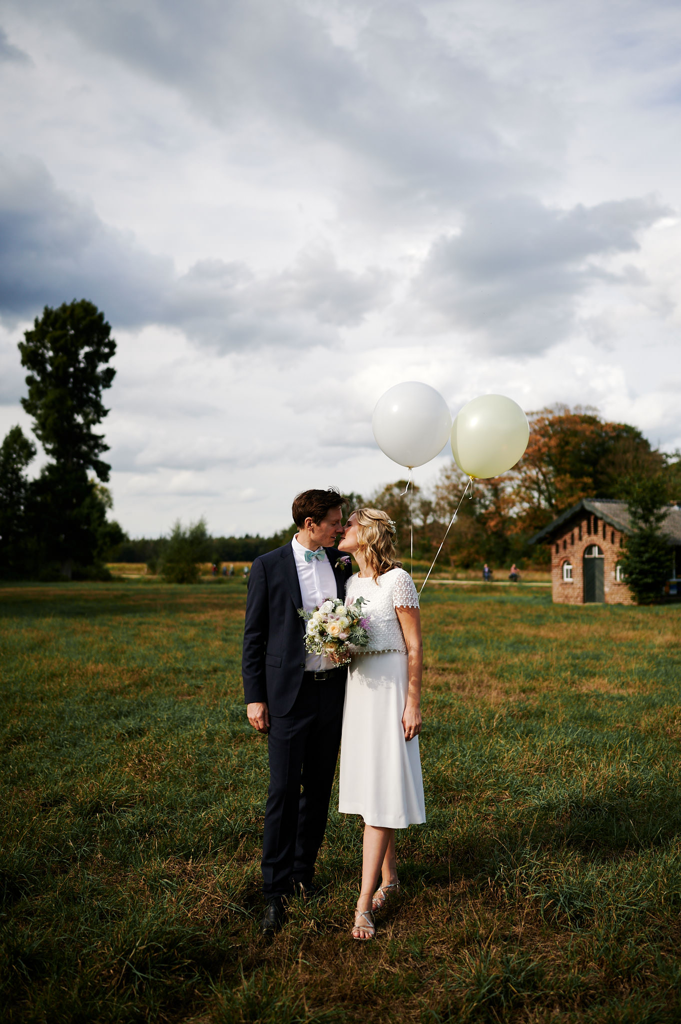 Ein Paar in Hochzeitskleidung hält Händchen und Luftballons auf einem Feld mit bewölktem Himmel darüber und feiert seine Hochzeit.