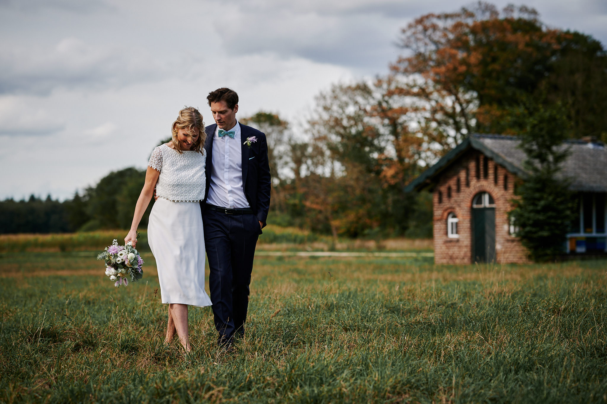 Ein Paar in Hochzeitskleidung geht durch ein Feld, die Braut hält einen Blumenstrauß und der Bräutigam legt seinen Arm um sie.