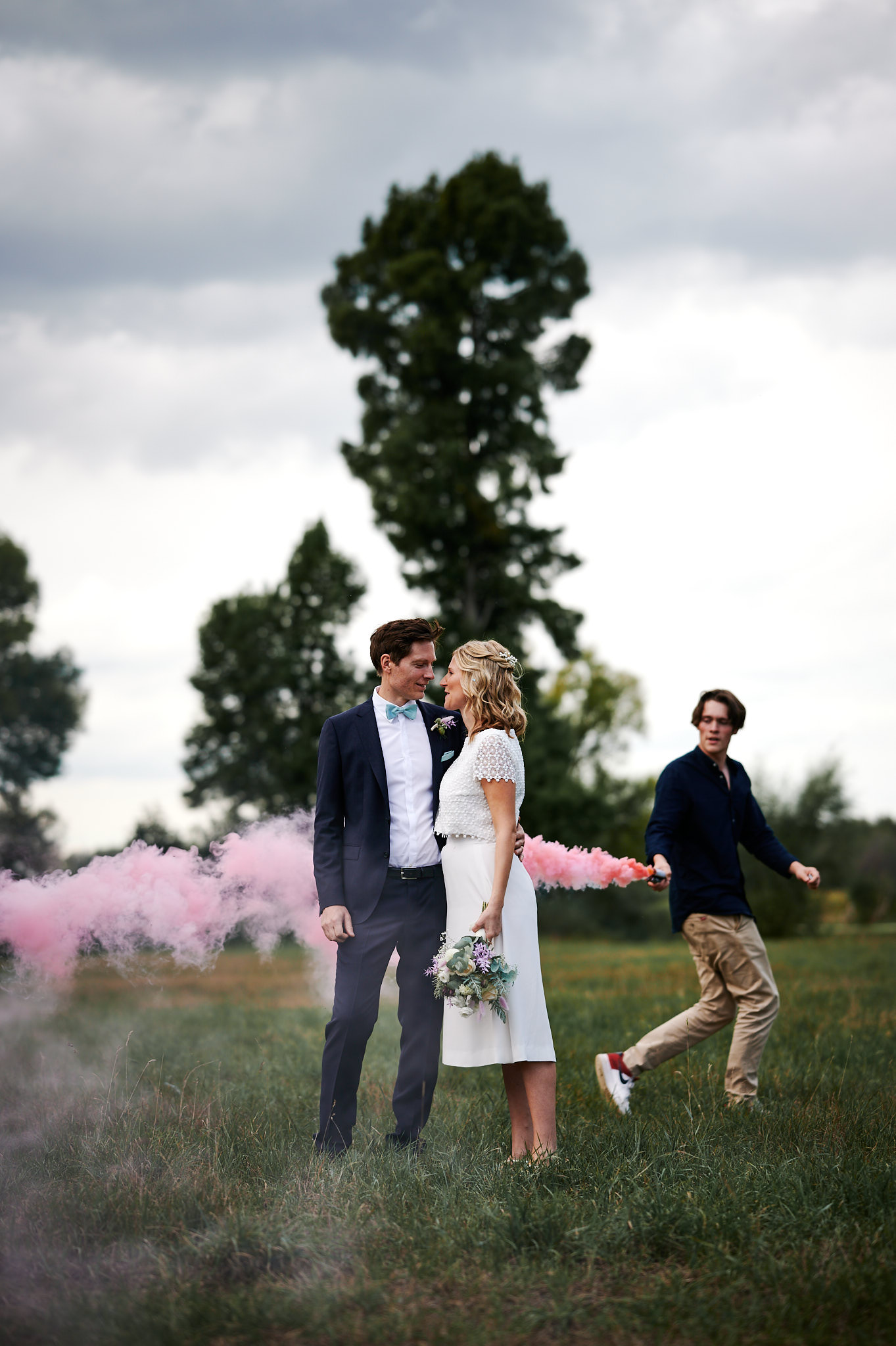 Ein Paar hält Händchen und schaut sich inmitten rosafarbenen Rauchs an, während eine andere Person im Hintergrund läuft, während ihrer Hochzeit auf Schloss Wissen.