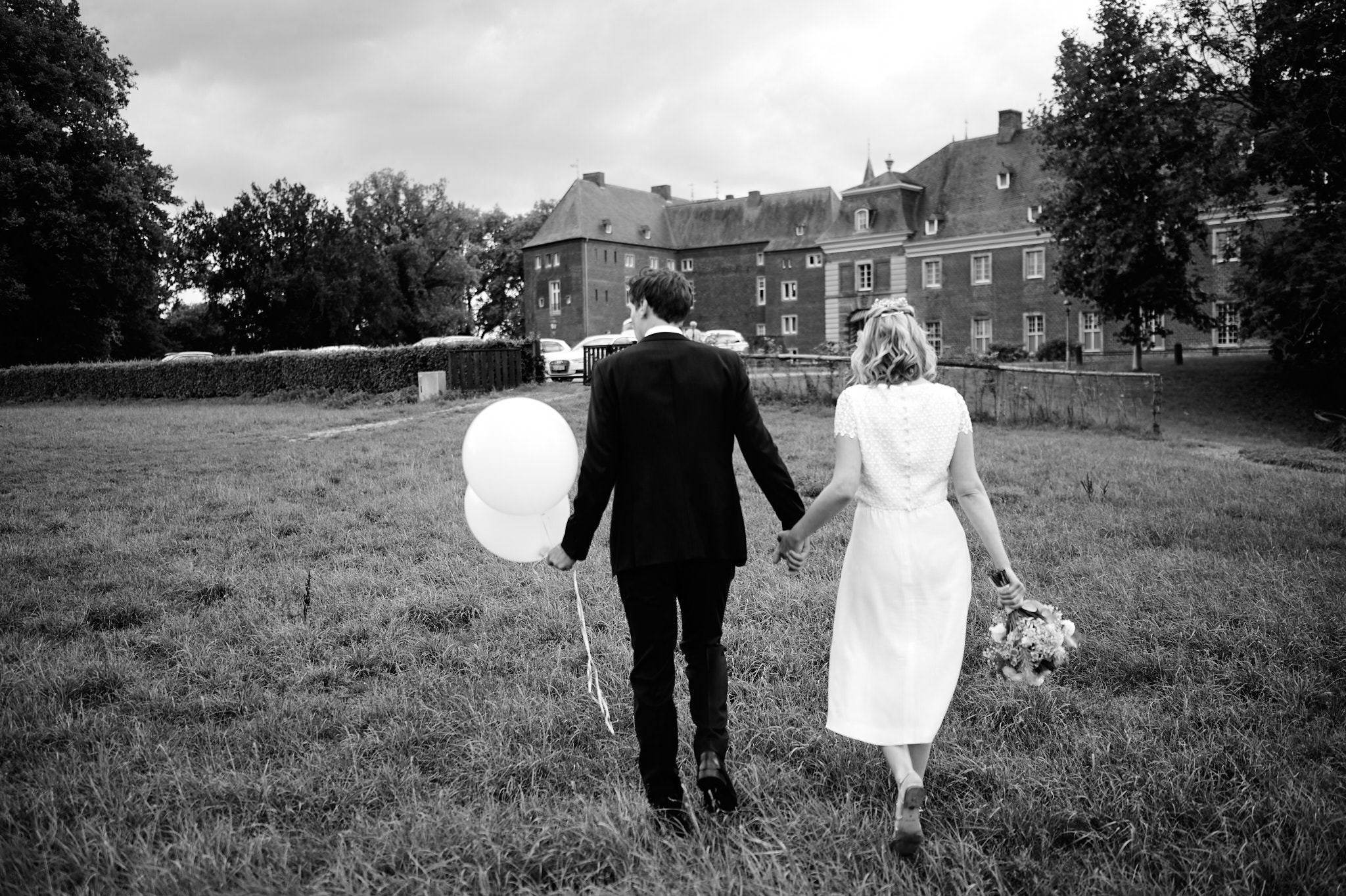 Händchenhaltend geht ein Paar auf Schloss Wissen zu, die Frau hält einen Blumenstrauß und Luftballons zur Hochzeit in der Hand.