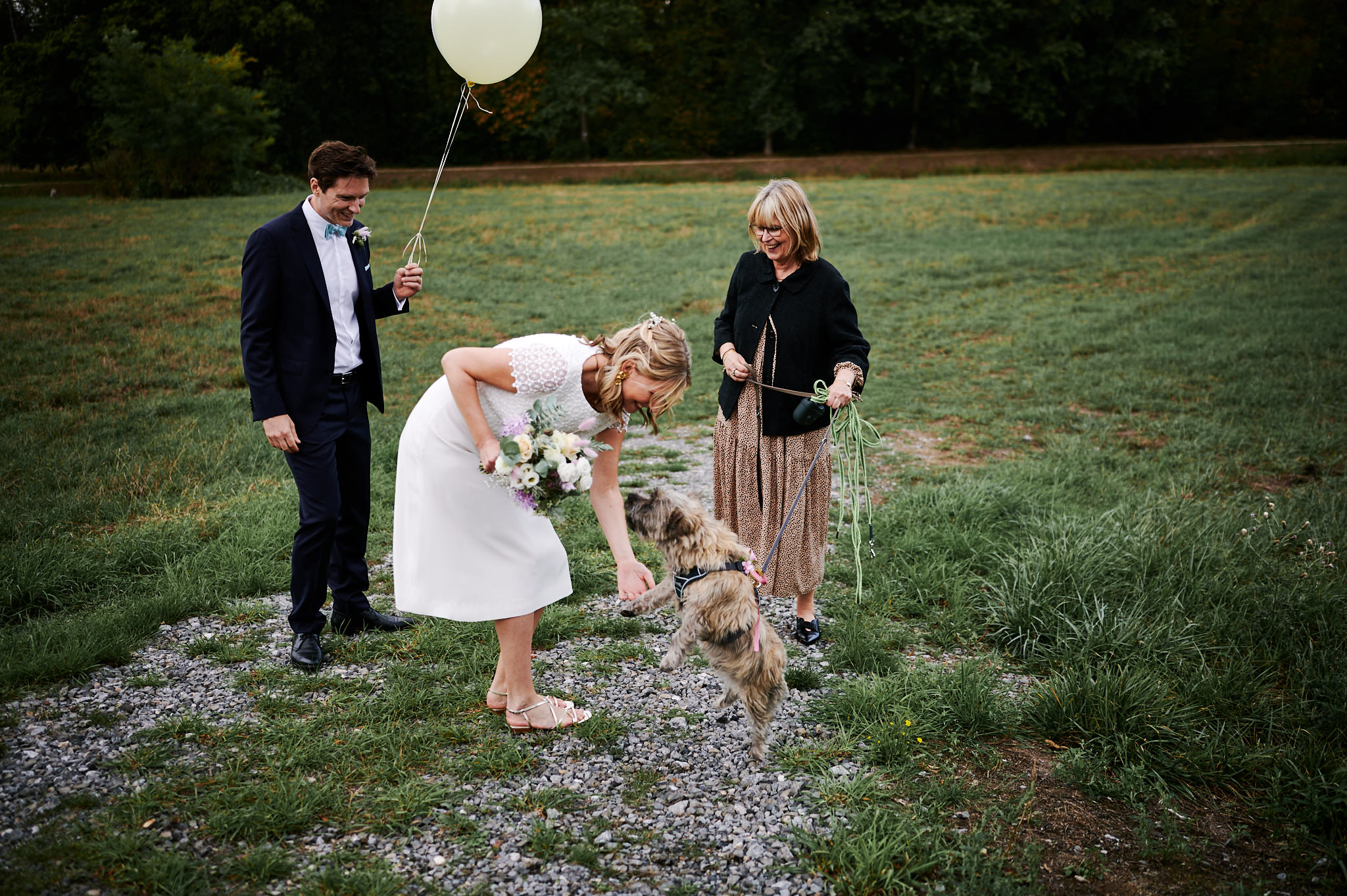 Braut streichelt einen Hund an der Leine, der von einer älteren Frau gehalten wird, daneben steht ein lächelnder Mann mit einem Luftballon im Freien auf dem Rasengelände von Schloss Wissen.