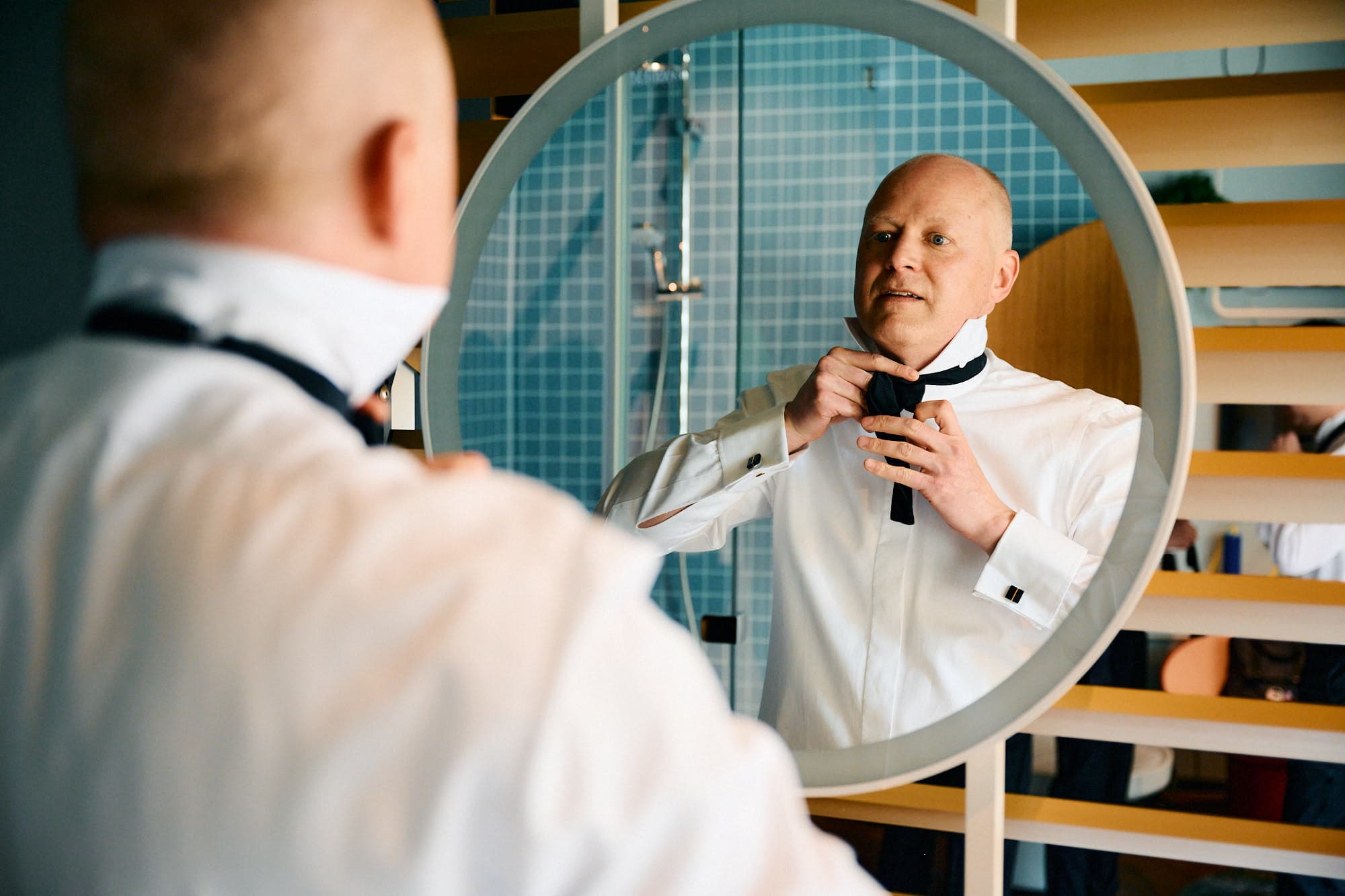 Ein Mann in einem weißen Hemd bindet seine Fliege, während er sein Spiegelbild im Badezimmerspiegel betrachtet und sich auf die Hochzeitsbilder vorbereitet.