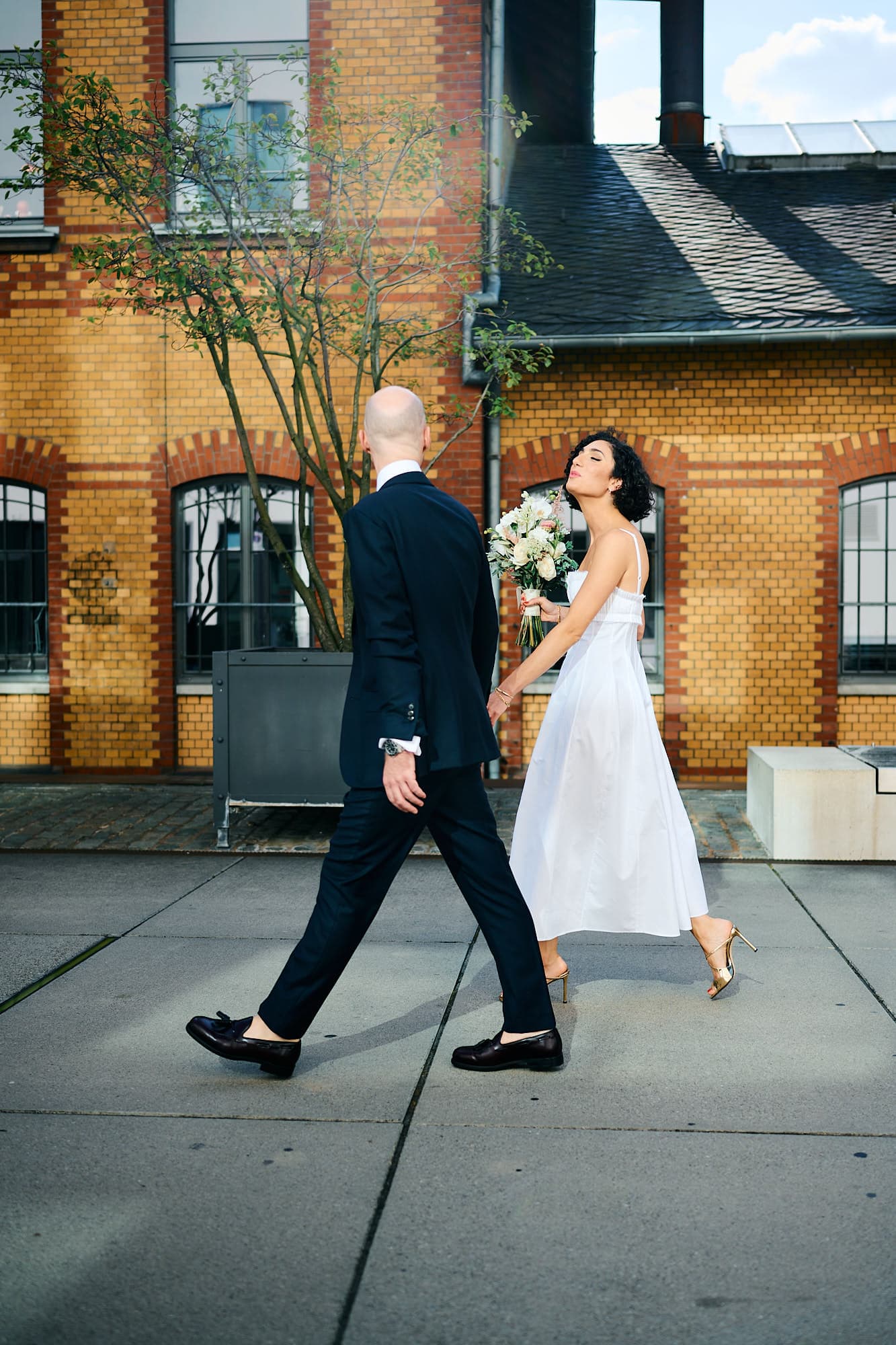 Ein Paar in formeller Kleidung geht auf dem Weg zu einer Hochzeits-Veranstaltung nach draußen, die Frau schaut ihr über die Schulter und der Mann geht voran.