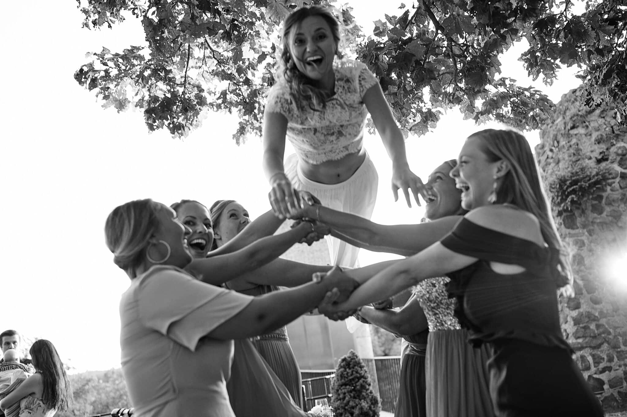 Eine Gruppe von Frauen hebt eine andere Frau in einer feierlichen Geste im Freien während Hochzeitsbildern in die Luft.