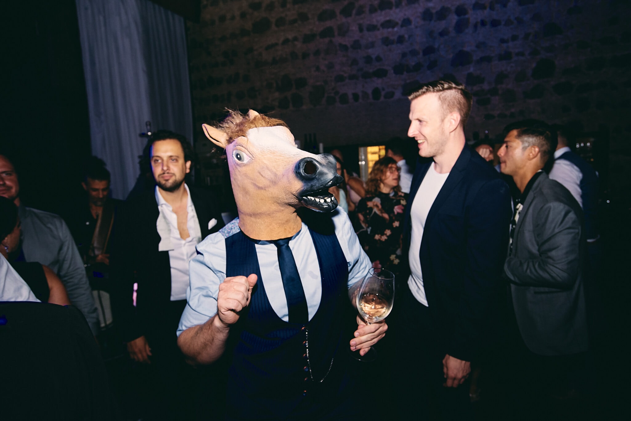 Eine Person mit Pferdekopfmaske hält bei einer Hochzeitsbilder-Veranstaltung einen Drink in der Hand, mit anderen Gästen im Hintergrund.