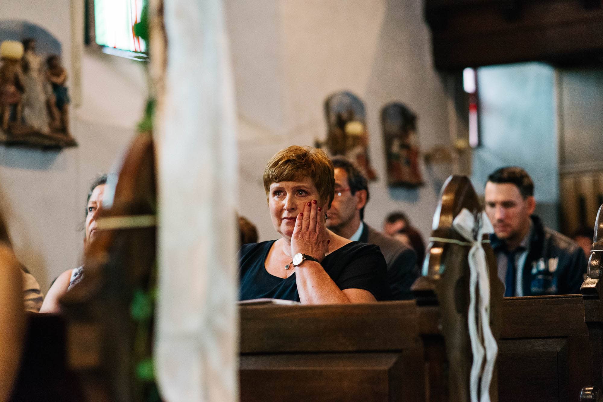 Frau sitzt in einer Kirchenbank, stützt ihr Gesicht auf ihre Hand und blickt zur Vorderseite der Kirche, während sie über Hochzeitsbilder nachdenkt.