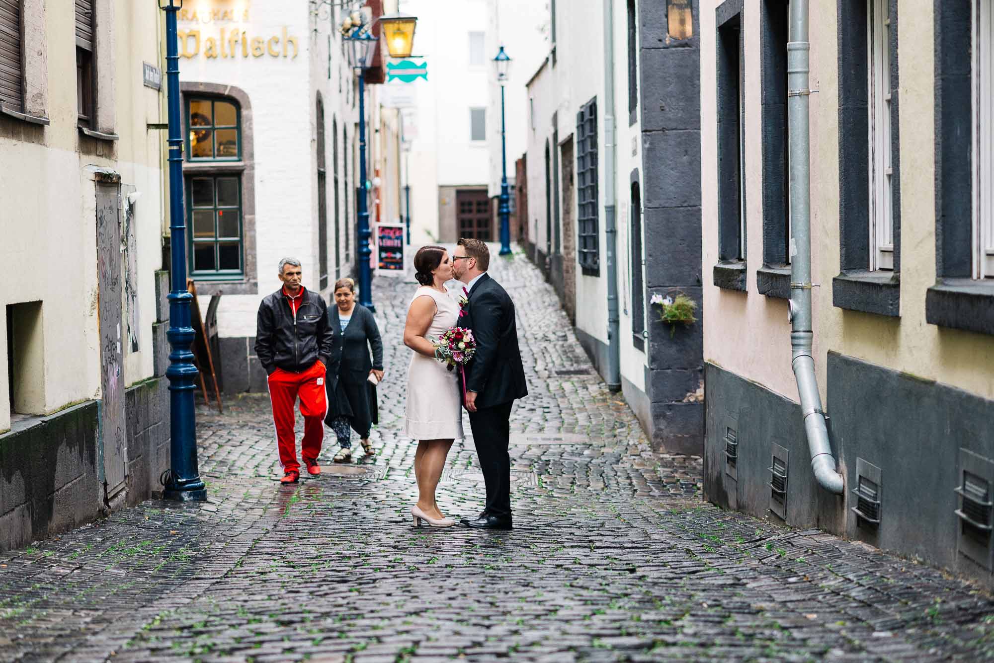 Ein Paar küsst sich auf einer Kopfsteinpflasterstraße, während ein Passant zuschaut und den Moment für ihre Hochzeitsbilder festhält.