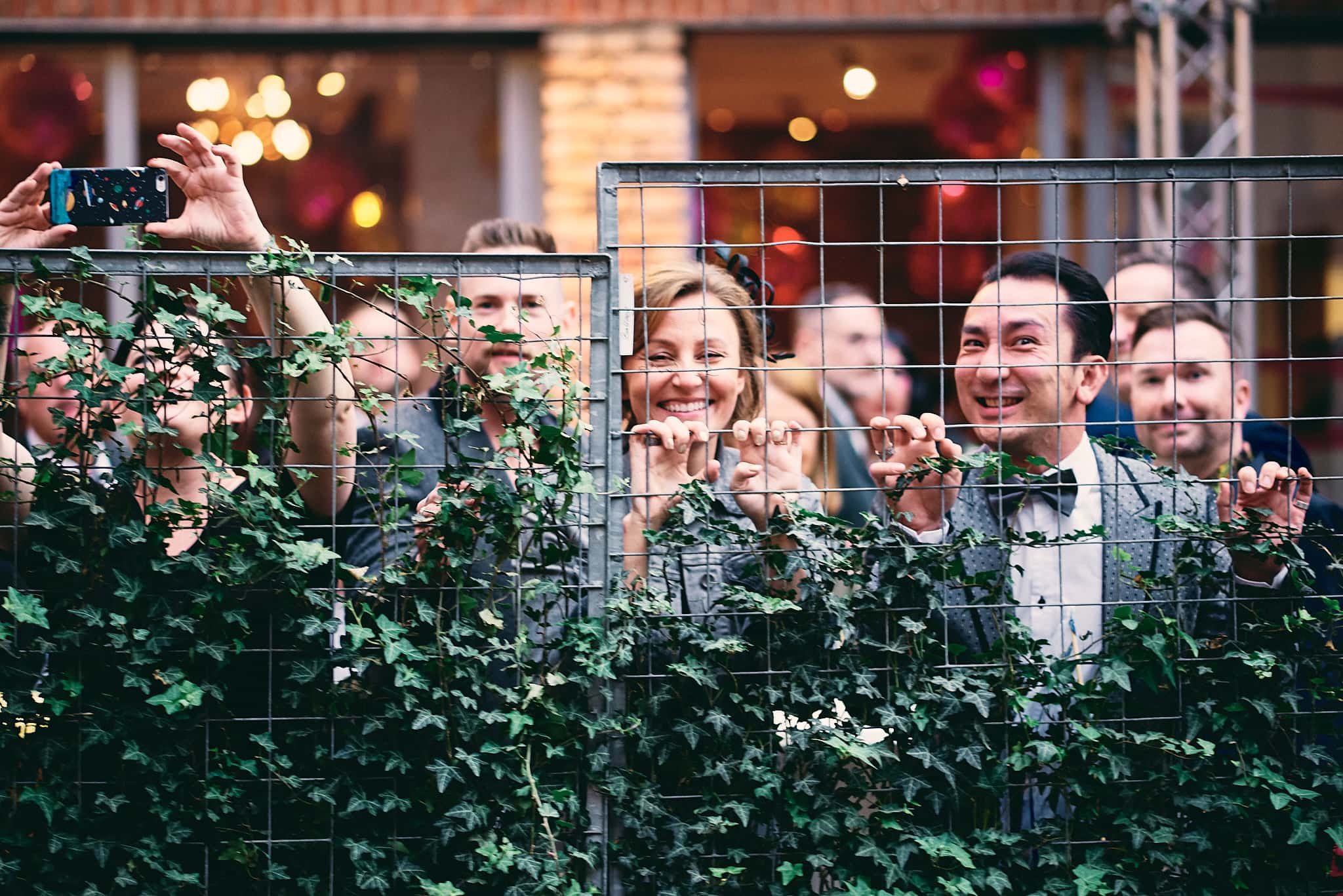 Menschenmenge hinter einem mit Grünpflanzen geschmückten Zaun, die Hochzeitsbilder mit dem Smartphone festhält.