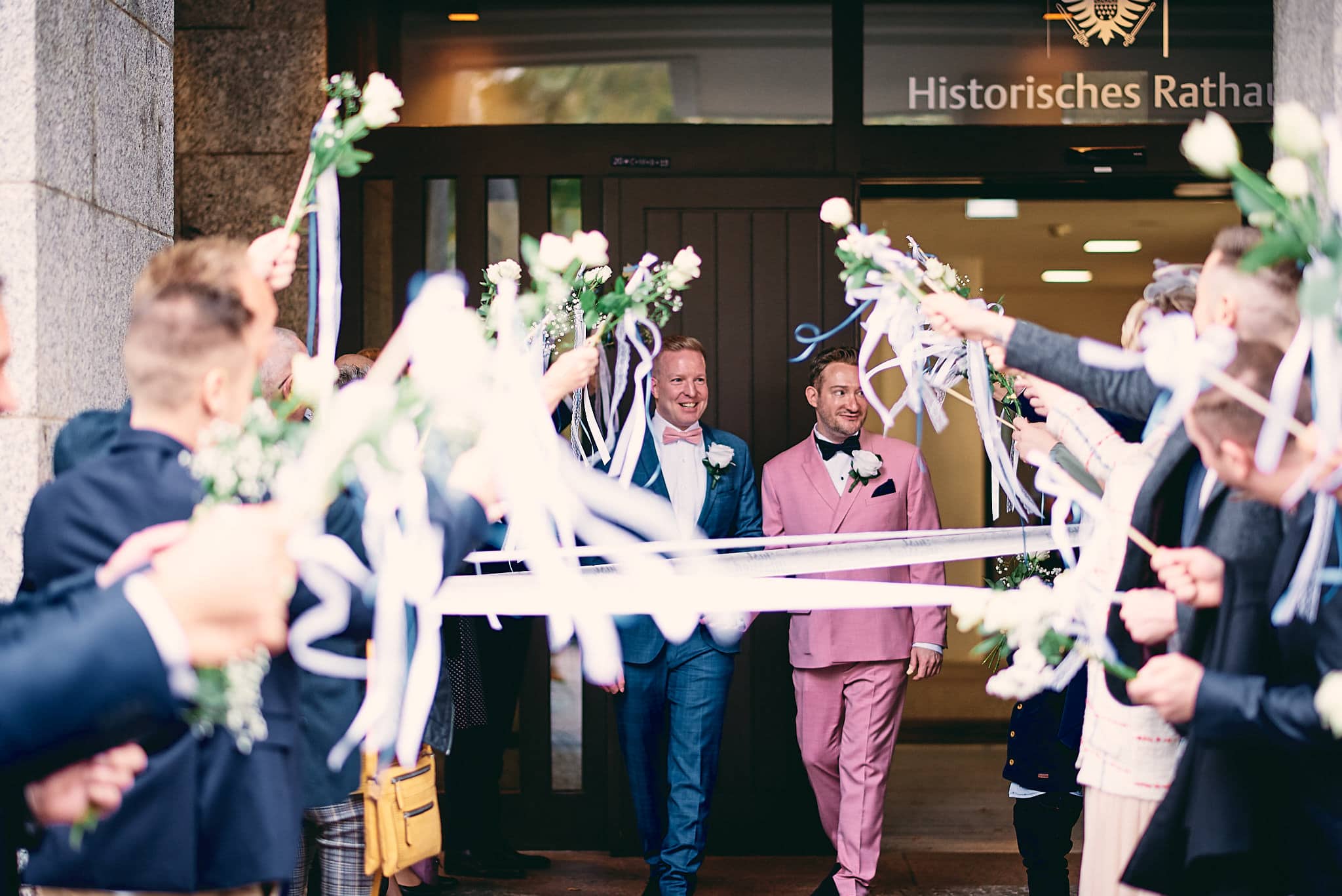 Ein fröhliches Paar geht durch einen Torbogen, begrüßt von Gästen vor dem historischen Rathaus und posiert für Hochzeitsbilder.