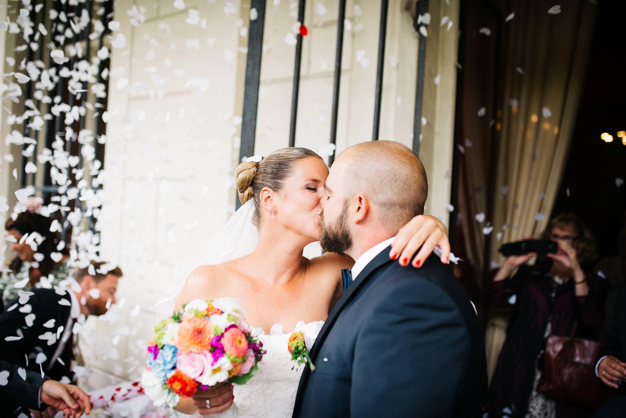 Braut und Bräutigam küssen sich inmitten von Konfetti vor dem Hochzeitsort für ihre Hochzeitsbilder.