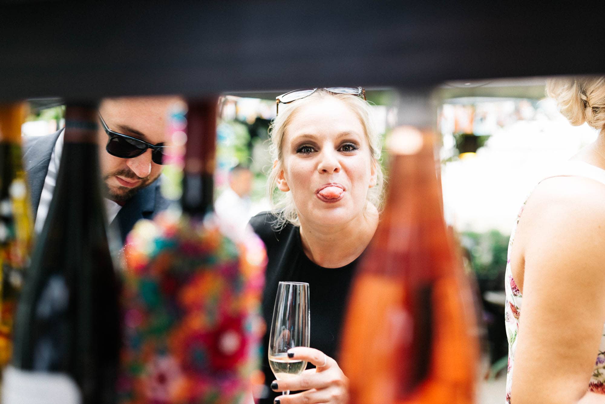 Frau streckt spielerisch die Zunge heraus, während sie ein Glas Wein in der Hand hält, im Hintergrund Regale mit Hochzeitsbildern und Flaschen.